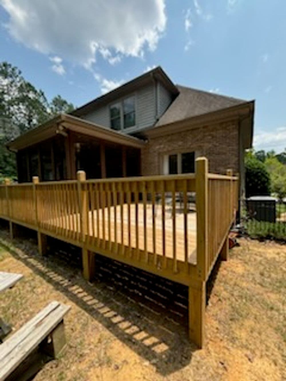Wooden deck attached to a brick home, surrounded by greenery under a blue sky.