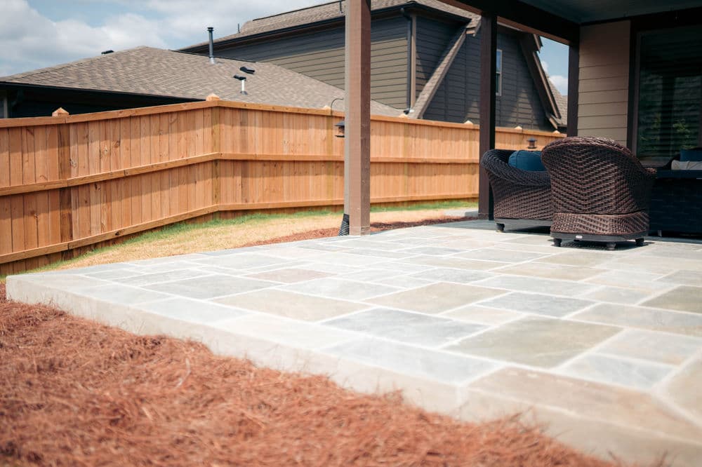 Patio area with stone flooring, wicker chairs, and fenced backyard in sunny outdoor setting.