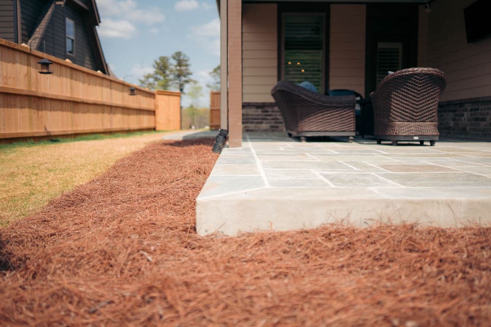 Patio with stone flooring, wicker chairs, and landscaped pine straw in a backyard setting.