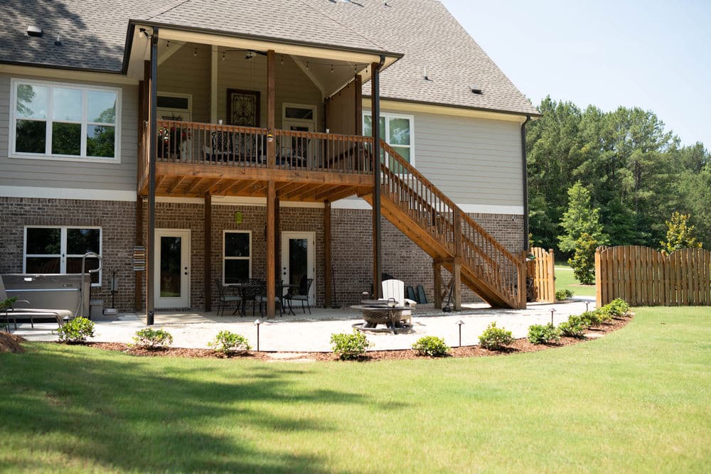 Two-story home with wooden deck, patio seating, and landscaped yard under clear sky.